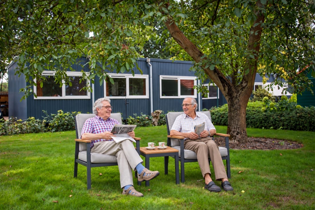 Bewoners zitten buiten bij hun tijdelijke mantelzorgwoning krant te lezen